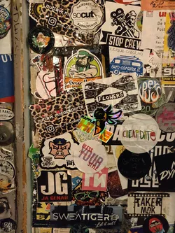 Street sticker Close-up view of a cluster of stickers, predominantly on a dark-colored surface.  The stickers vary in shape, size, design, and color.  One prominent sticker displays a holographic or iridescent effect. Some stickers feature text, logos, and illustrations.