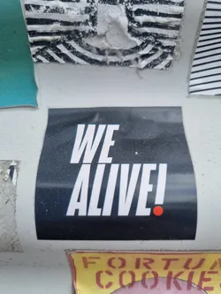 A black sticker with the text "WE ALIVE!" in white bold capital letters. There is a small red dot at the end of the text. 