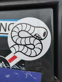 Street sticker A round sticker featuring a simple line drawing of a worm. The worm is depicted in black and white on a white background, with the worm's body segmented and curled around itself. The sticker appears to be placed on a dark gray or black surface, and other stickers are visible in the background. 
