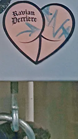 Street sticker The sticker is shaped like a heart and is divided into two symmetrical sections resembling a pair of buttocks. The text 'Ravian Derriere' is written in a gothic-style font. There are also stylized arrows pointing towards the center of each 'cheek'.