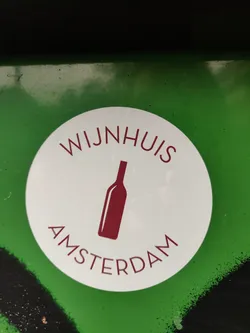 Street sticker Round white sticker with a wine bottle icon and the text 'WIJNHUIS AMSTERDAM'. The sticker is on a green surface.