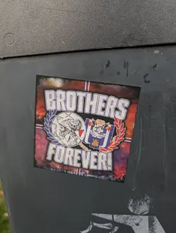 Street sticker The sticker features a central emblem with two distinct halves. The left side depicts a profile of a bearded man wearing a helmet, rendered in grayscale, surrounded by a laurel wreath. The right side displays a crest with a crown, flags, and a golden lion, all in shades of blue and red. The text 'BROTHERS FOREVER!' is prominently displayed in large, stylized white letters with a purple outline, arching over and under the central emblem. The background is a distressed, fiery red and purple gradient, giving it a worn and intense aesthetic. Small flags, possibly representing national or club affiliations, are visible within the emblem.