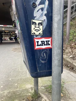 The sticker at the center of the image is a well-known 'Obey Giant' design, featuring a stylized black and white face with large eyes and an open mouth. It is placed on a dark blue, possibly metallic, surface. Below the Obey Giant sticker is another sticker with a red border and a white background, displaying the bold black letters 'LRK'. The overall presentation suggests urban art or graffiti culture.