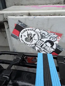 The sticker features two distinct designs layered on top of each other. The top sticker has a camouflage-patterned background with bold white lettering that appears to read 'DAS'. Below this, a circular sticker is visible. The circular sticker has a black and red striped border. Inside, it depicts a winged cherubic figure, possibly a lion, standing on a globe or a detailed base, with the text 'TURN-U-SPIELVEREIN LEVERKUSEN' and '19 04' also visible. Partially overlapping this circular sticker is another element with a stylized face shouting, with the words 'ULTRA' and 'LEVERKUSEN' repeated. The overall impression is one of fan culture and sports team affiliation.