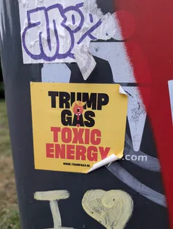 Street sticker The sticker is rectangular with a bright yellow background. Dominating the center is bold black text reading 'TRUMP GAS' with a small, stylized caricature of Donald Trump, depicted with his signature hair, a red tie, and an open mouth, positioned between 'TRUMP' and 'GAS'. Below this, in large red capital letters, are the words 'TOXIC ENERGY'. At the bottom of the sticker is a website address: 'WWW.TRUMPGAS.NL'. The sticker appears to be slightly creased and peeling at the edges, indicating it has been adhered to a surface.