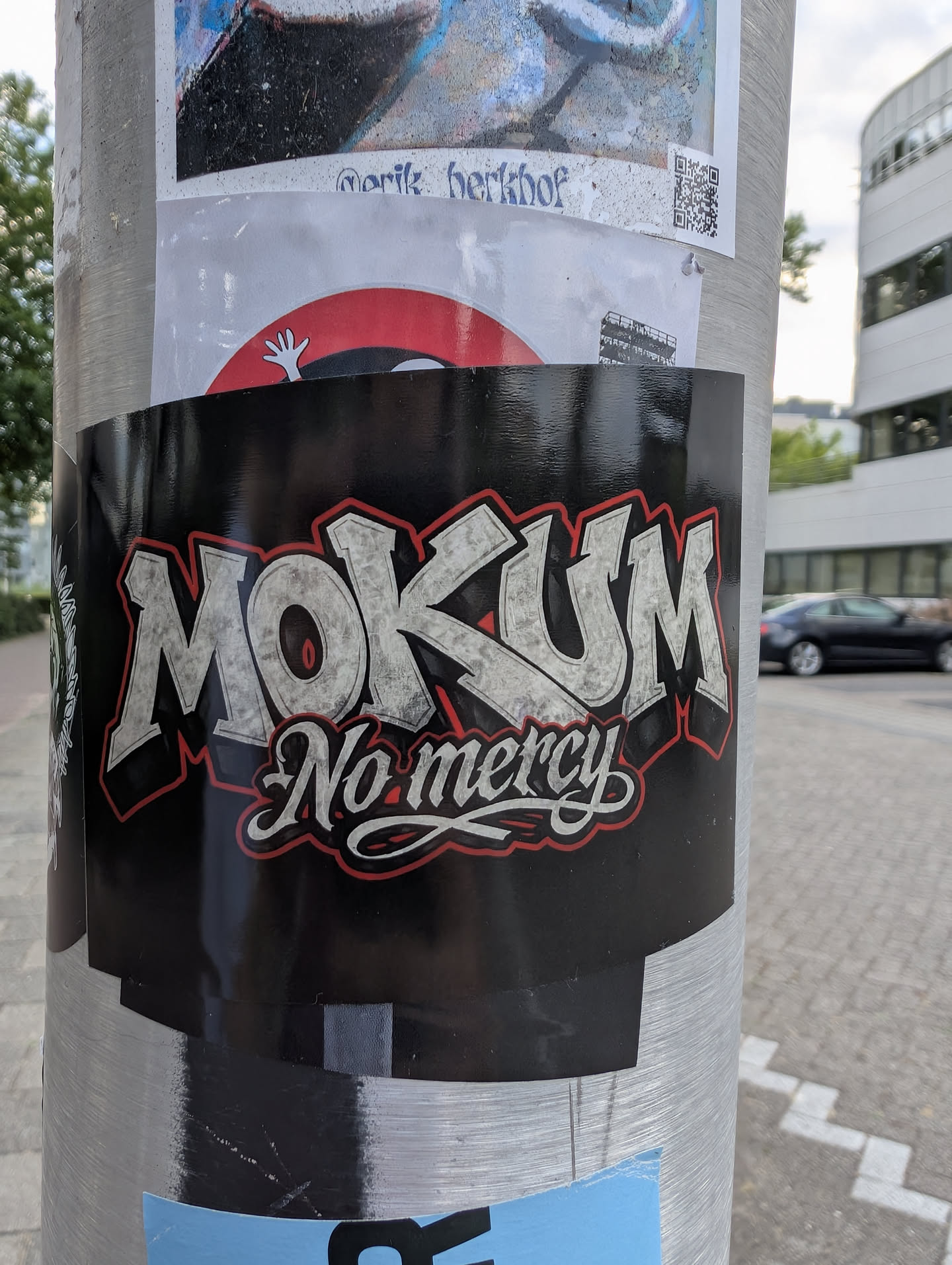 A rectangular black sticker featuring the word 'MOKUM' in large, textured silver block letters with a red outline. Below, 'No mercy' is written in a white cursive script, also outlined in red.