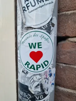 Street sticker A round sticker with a green border and a red heart in the center. The text "Freunde des Spaβes" is written in a cursive font above "WE", and "RAPID" is written in a larger font below the heart. 