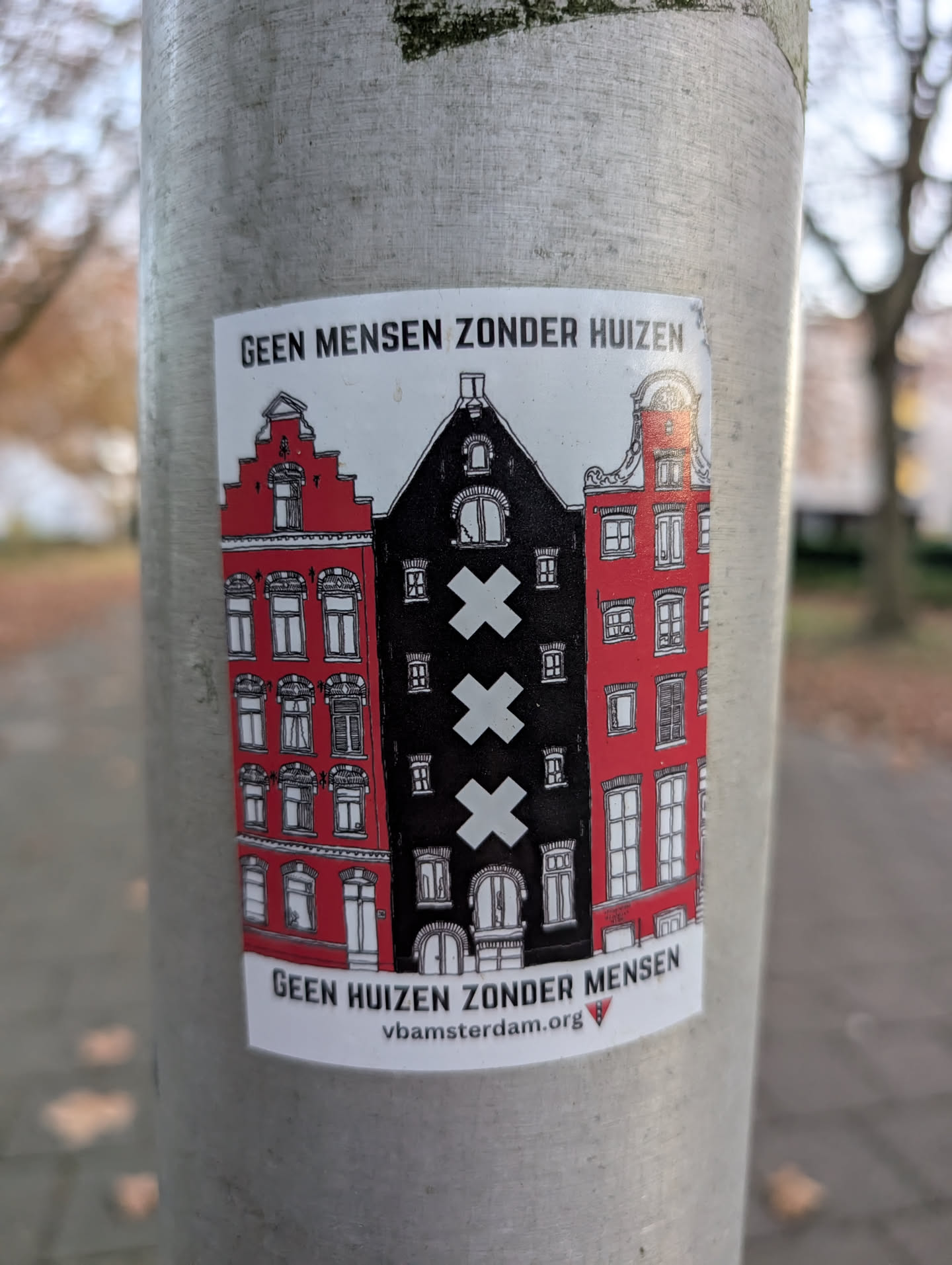 The sticker features a stylized depiction of three Dutch canal houses. The two outer houses are bright red with white window frames and intricate architectural details, including stepped gables. The central house is black and features the three St. Andrew's crosses, a prominent symbol of Amsterdam, in white. The houses are rendered in a graphic, somewhat simplified style with black outlines. The sticker has a white background and is affixed to a metal pole.