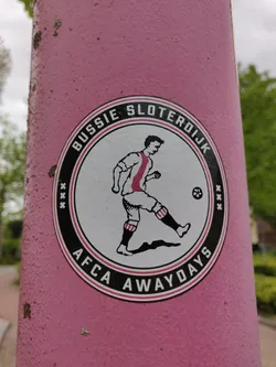 Street sticker A round sticker featuring a football player in vintage attire. The text around the image says 'Bussie Sloterdijk' on the top and 'AFCA Awaydays' on the bottom. The sticker is affixed to a pink pole.