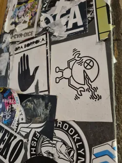 Street sticker Close-up of a sticker featuring a simple, graphic design. The sticker is black and white and depicts a small figure that appears to be holding or interacting with a slice of pizza. The style is minimalistic and geometric.