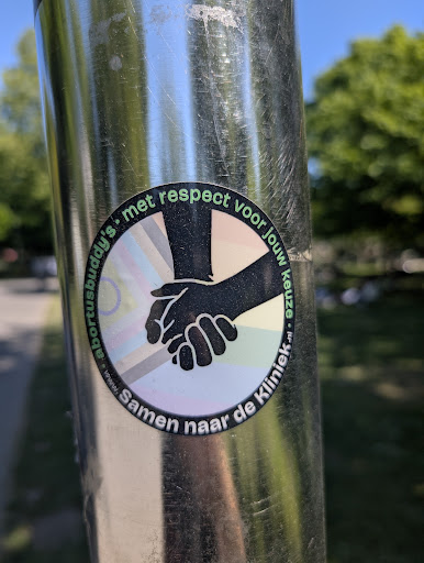 The sticker on the pole features two hands clasped together in a supportive gesture. The text 'met respect voor jouw keuze' (with respect for your choice) arcs above the hands, and 'Samen naar de Kliniek' (Together to the Clinic) arcs below. The sticker promotes respect for personal choice and suggests support for people visiting clinics.