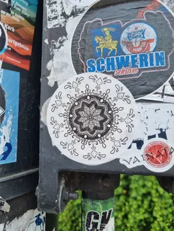 Street sticker A round sticker with a detailed mandala design in black and white. The mandala is surrounded by a border of stylized droplets. The sticker is affixed to a dark gray pole, which also has other stickers on it.