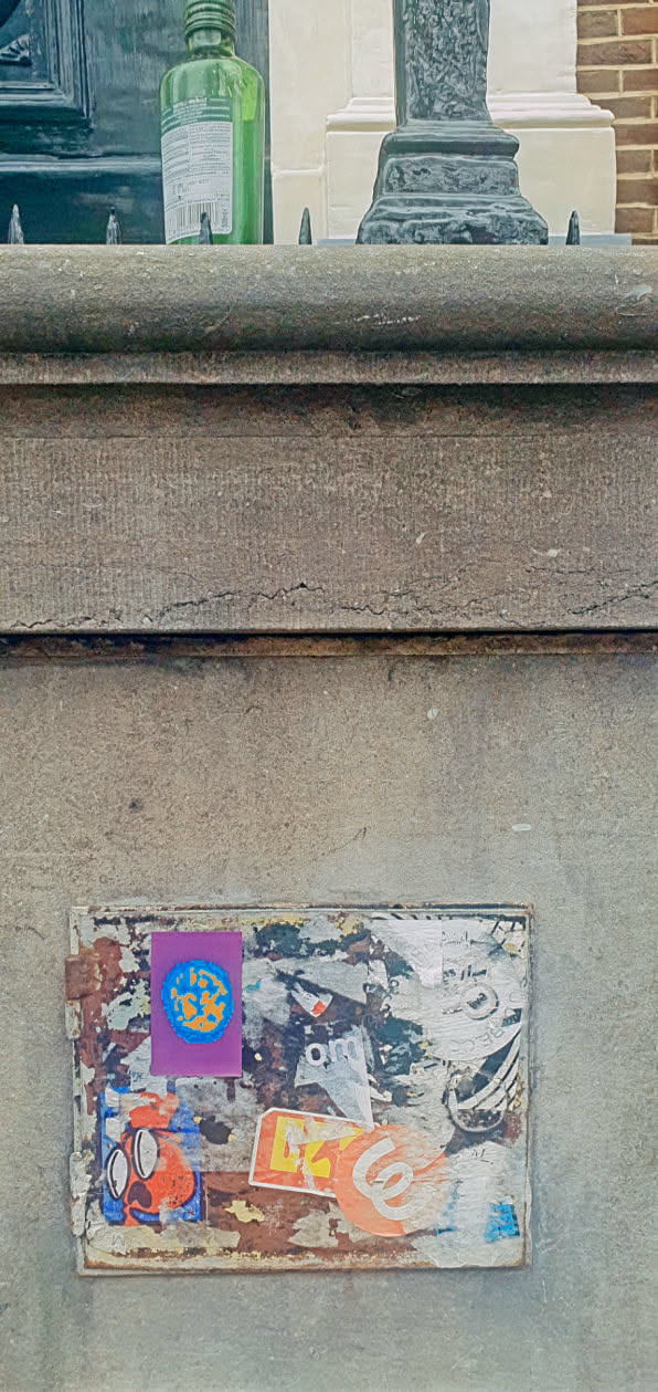 This image features a section of a textured, likely aged concrete surface, adorned with multiple layers of stickers. The primary focus is a rectangular sticker area covered with diverse images and shapes. Most notable is a purple sticker at the top left corner displaying an earth-like design. Various other stickers with geometric patterns and faded colors are scattered around it. The stickers appear faded and peeling, indicative of sun exposure and time.