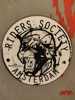 Round sticker featuring a black and white illustration of a snarling panther's head wearing a helmet. The text "RIDERS SOCIETY AMSTERDAM" encircles the illustration. A small logo is visible at the bottom left.