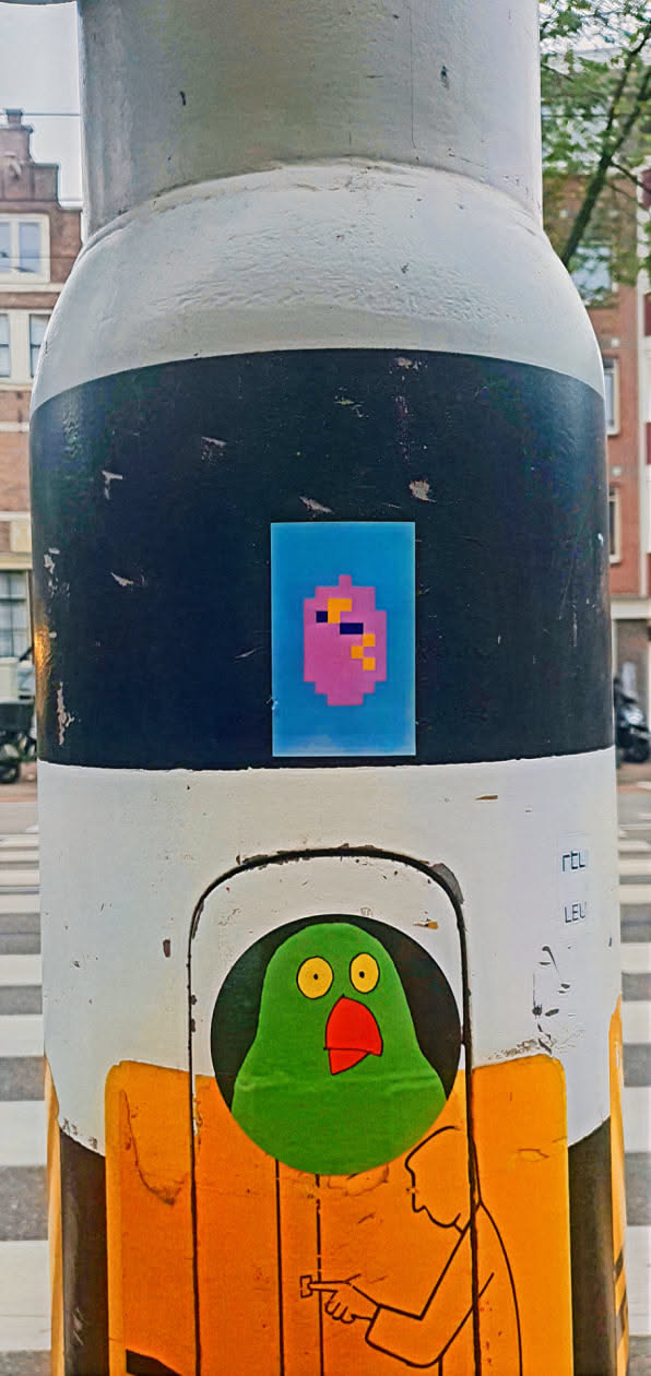 A sticker featuring a pixelated figure with a pink face, dark blue eye, and orange pixels on top of its head. The sticker is placed on a pole with a black stripe. Below it, another sticker with a green bird and an illustration of a person can be seen.
