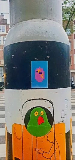 Street sticker A sticker featuring a pixelated figure with a pink face, dark blue eye, and orange pixels on top of its head. The sticker is placed on a pole with a black stripe. Below it, another sticker with a green bird and an illustration of a person can be seen.