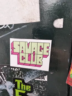 Street sticker A white square sticker features the words "SAVAGE CLUB" stacked on two lines. The text is rendered in a light green fill with a bold pink outline, giving a three-dimensional, retro vibe. The sticker is pasted on a weathered surface, with other stickers and graffiti visible around it.