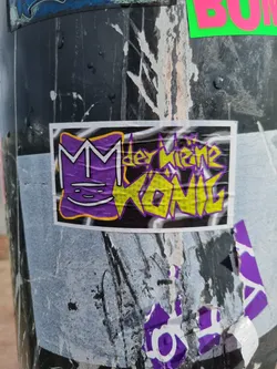 Street sticker A sticker featuring a stylized 'M' with a crown and the text "Der kleine König" (The little king) in graffiti-style lettering. The sticker is purple and yellow on a black background. 