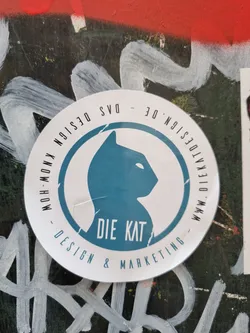 A round sticker with a teal cat silhouette and the text "Die Kat" in the center. The outer circle says "www.die-kat.design -  muh-meow - design & marketing".