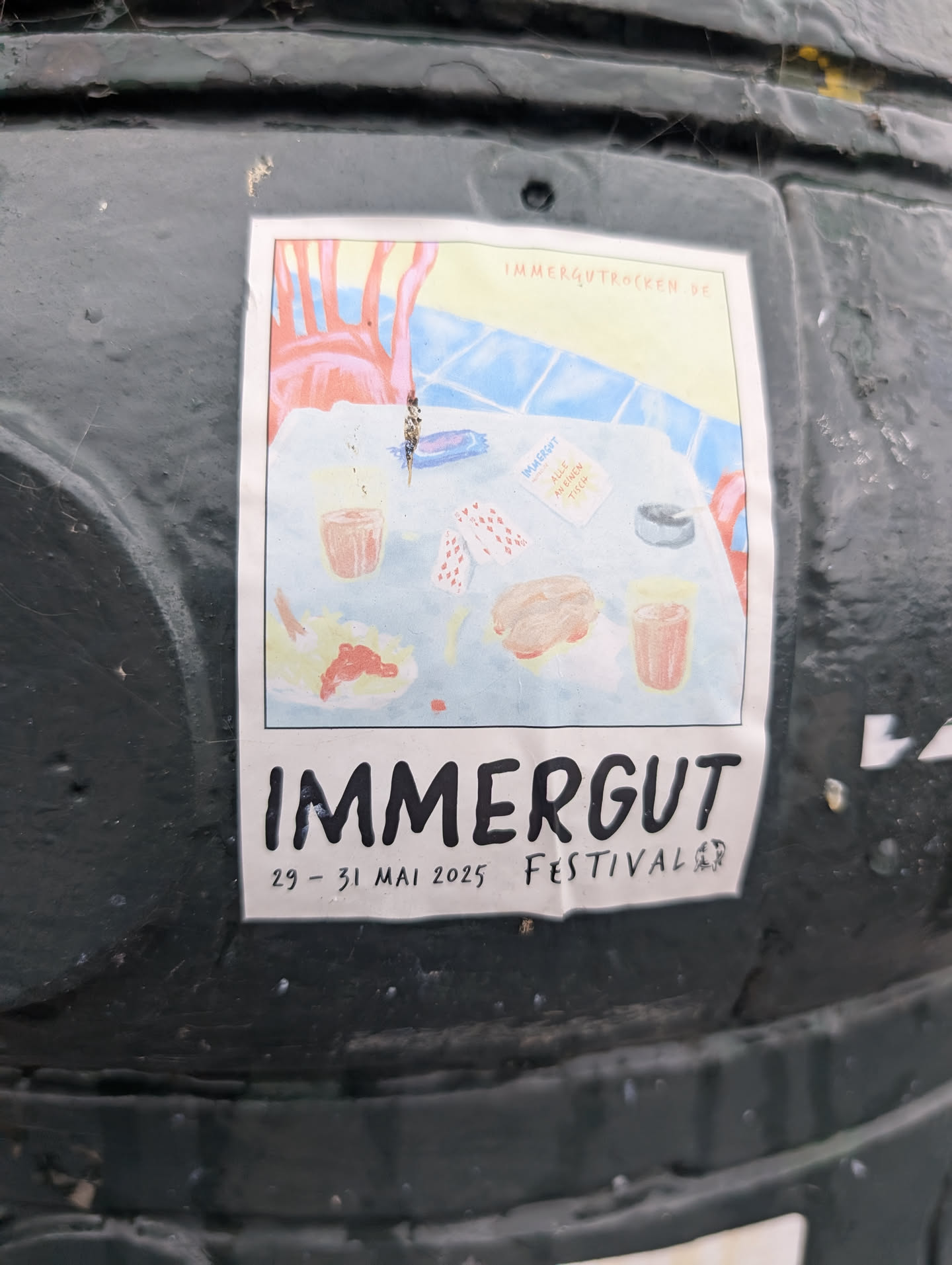 A sticker promoting the IMMERGUT FESTIVAL, scheduled for May 29-31, 2025. The central graphic is a whimsical illustration of a table setting with two glasses of drinks, a burger, and some playing cards. The background depicts a stylized scene with red chairs and a patterned surface. The text 'IMMERGUT' is prominently displayed in large, bold, black letters at the bottom, followed by the dates and 'FESTIVAL'. There is also a smaller text box on the illustration that reads 'IMMERGUT ALLE AN EINEN TISCH' and a website address 'IMMERGUTROCKEN.DE'.