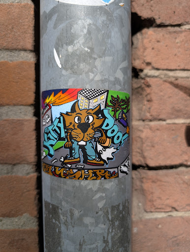 This sticker features a cartoon cat character in jeans and shoes, surrounded by graffiti-style elements, including buildings, a tree, and dog bones. The background includes a building with a checkered roof and some graffiti, adding to its urban art style. The sticker is attached to a metal pole.