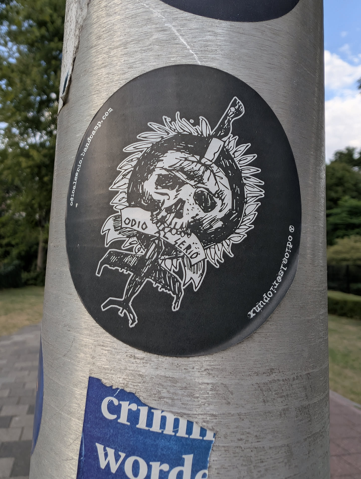 An oval black sticker with a white graphic of a skull impaled by a dagger. A banner across the skull reads 'ODIO AL SERIO'. The design is framed by a spiky circular border, with band links printed along the sides.