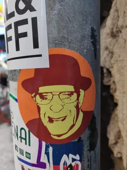 Street sticker This round sticker showcases a vibrant, almost pop-art-like portrait of a smiling man. He's depicted wearing a fedora and glasses, giving him a classic, retro feel. The color palette is bold and striking, with the main image in shades of orange, yellow, and red against a contrasting background. The sticker's style is simple yet eye-catching, perfect for adding a touch of personality to any surface.