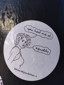 Street sticker A round sticker with a line drawing of a woman and speech bubbles. One speech bubble says "you had me at..." and the other says "equality".  The text "lekker-blijvenlikken" is written at the bottom. 