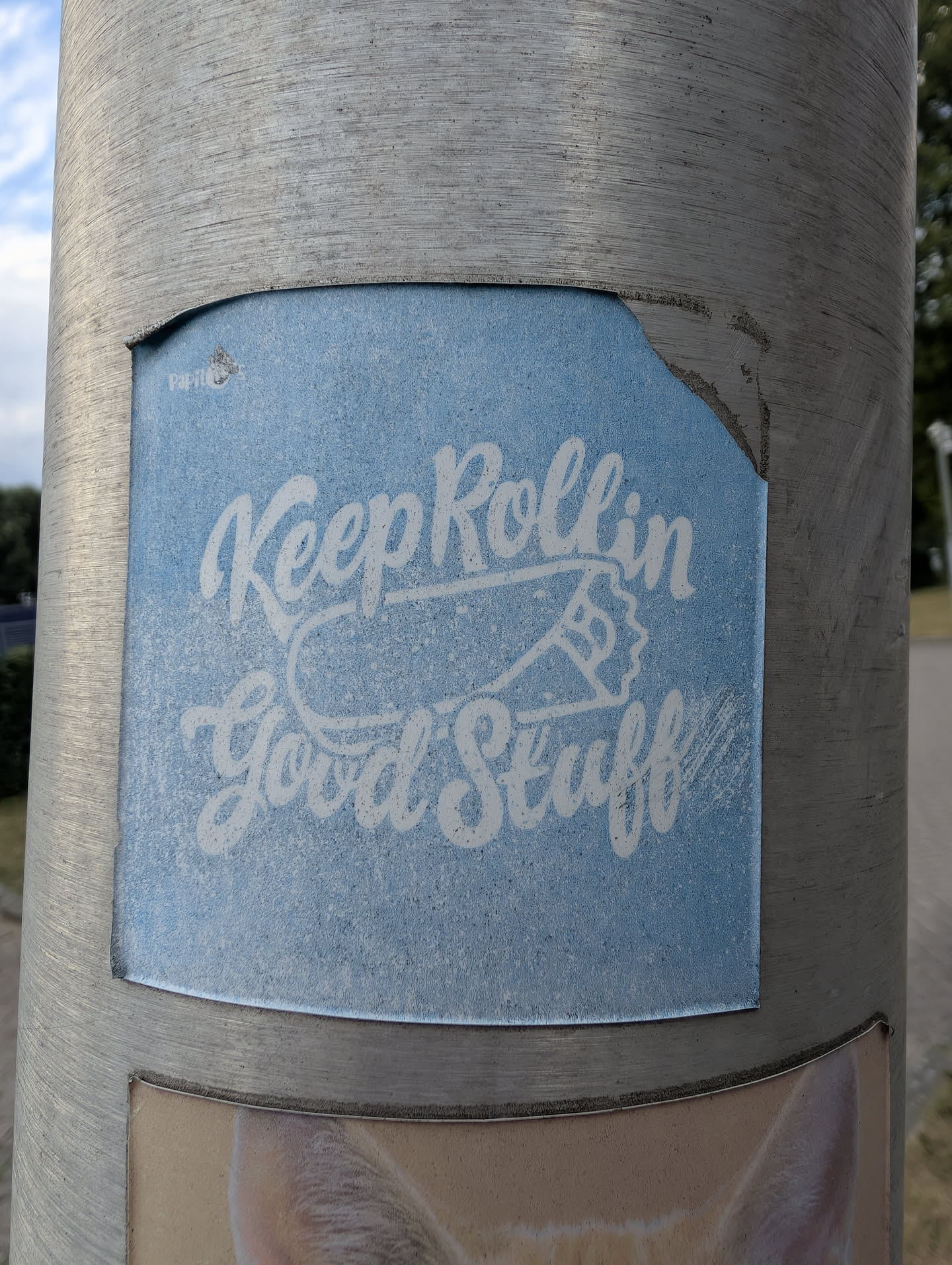 A square light blue sticker on a metal pole. It features white cursive text 'Keep Rollin Good Stuff' surrounding a simple line illustration of a rolled joint. A small 'Papito' logo is in the top-left corner, and the top-right corner is torn.