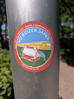 A round sticker featuring a bus and a train is stuck on a metal pole. The sticker's text reads "Wij Reizen Samen" and mentions a website. The sticker promotes good working conditions for public transport workers.