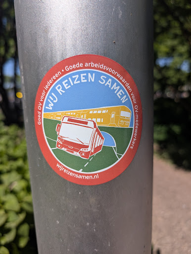 A round sticker featuring a bus and a train is stuck on a metal pole. The sticker's text reads "Wij Reizen Samen" and mentions a website. The sticker promotes good working conditions for public transport workers.