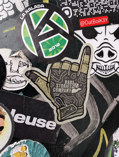 This sticker for 'Bad Stories Company' shows a hand gesture, specifically the 'rock on' or 'devil horns' sign, rendered in a topographic map style with earthy tones. The company's name is printed within the hand's palm area.