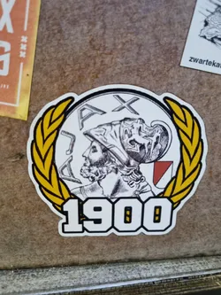 A sticker featuring the Ajax Amsterdam logo, which includes a laurel wreath, the year 1900, and a depiction of a warrior's head. The style is vintage and classic.