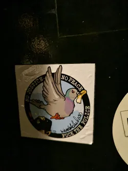 A sticker featuring a cartoon pigeon carrying a piece of paper in its beak. The text around the pigeon says "No Justice, No Peace" and "Fuck the Police." There's also a signature that reads "Wandelclub030."