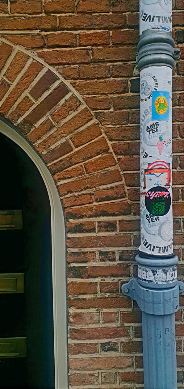 A grey pole is covered in stickers and is situated next to a brick wall and doorway. The stickers have various designs and logos. Some visible stickers include “AMS TEF DAM LIVE”, “YouTube”, “Z06”, and “CYBER ATELIER”.