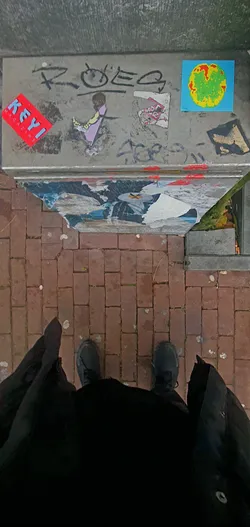 A collection of stickers, graffiti and torn posters are adhered to a metal surface. The leftmost sticker has red background and white text. Some of the stickers are partially damaged. Some graffiti is seen on top of the stickers.