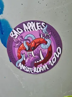 Street sticker Round sticker featuring a cartoon illustration of a monstrous apple infested with worms. The style is playful and slightly grotesque. The text 'Bad Apples' is prominently displayed, along with 'Amsterdam 2020' at the bottom. The artist's signature is visible on the right side of the sticker.