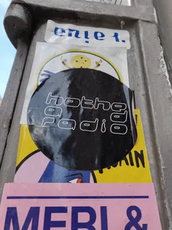 Black round sticker with the text "hotheya radio" in a stylized font.  It's stuck on a pole alongside other stickers.