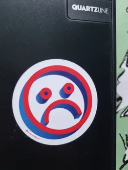 A round sticker with a stylized sad face in red, white and blue. The face is designed in a 3D style with a thick border. The sticker is placed on a black surface.  It includes the social media handle @Laadio.