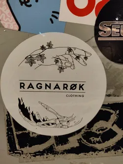 Street sticker Round white sticker with black line art depicting a hand intertwined with leaves on one side and a skeleton hand engulfed in flames on the other.  The text "RAGNAROK" and "CLOTHING" is visible in the center.