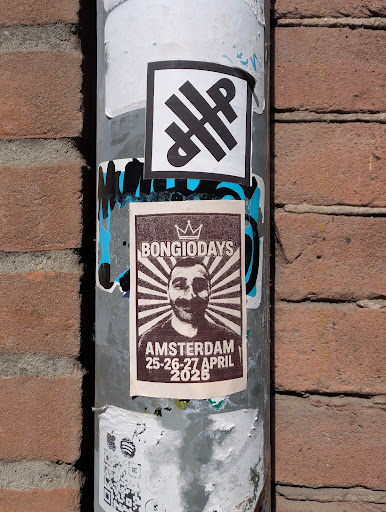 A poster on a pole advertises "Bongiodays" in Amsterdam from April 25-27, 2025. The poster features a portrait of a man with a mustache. There's another sticker with an abstract black design above the poster and a QR code sticker below it.