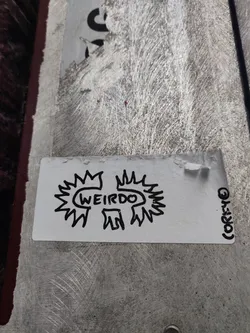 Street sticker A white sticker with a hand-drawn design. The design features the word 'WEIRDO' enclosed in a sun-like shape with spiky rays. There's a small, barely visible signature or logo in the bottom right corner. The sticker is affixed to a textured surface, possibly metal or concrete.