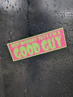 Street sticker A rectangular sticker with a bright pink background and neon green text. The text is in a bold, blocky font. The top line reads 'BUT MOMMY SAYS I'M A' and the bottom, larger line reads 'GOOD GUY'. The sticker is slightly angled and appears to be adhered to a dark, textured surface covered in water droplets.