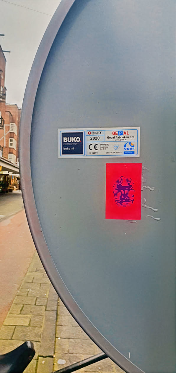 A red square sticker with a pixelated purple design is affixed to a gray, slightly curved surface. The surface appears to be part of a larger structure. Next to the sticker, there's a technical label that reads 'BUKO INFRASUPPORT', 'GEPAL' and 'VNVF'.