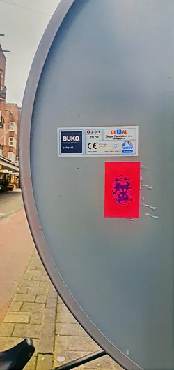 Street sticker A red square sticker with a pixelated purple design is affixed to a gray, slightly curved surface. The surface appears to be part of a larger structure. Next to the sticker, there's a technical label that reads 'BUKO INFRASUPPORT', 'GEPAL' and 'VNVF'.