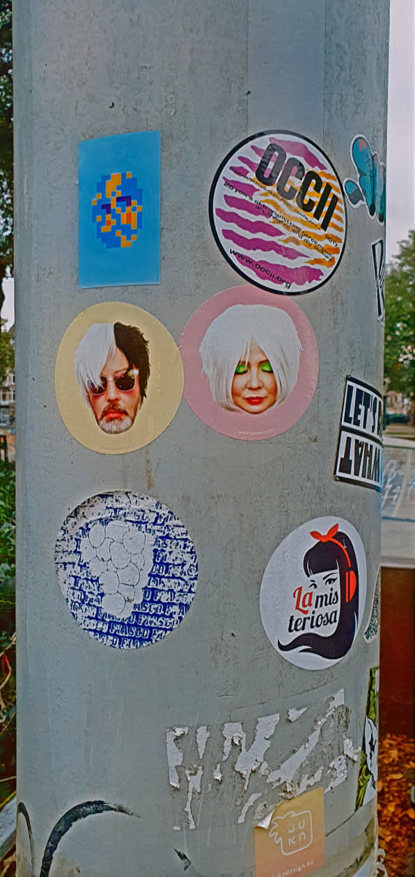 This image shows several stickers adhered to a pole. The stickers include a pixelated design on a blue background, a round sticker with a man's face split between black and white hair, a round sticker with a woman's face and white hair on a pink background, a sticker with the text "La misteriosa" and an illustration of a woman with headphones, and a sticker for OCCII with its website address.