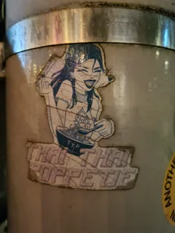 Street sticker A sticker featuring an illustration of a manga-style girl happily eating a bowl of noodles. The sticker has a worn look, with some visible cracks and tears. The text "Thai Thai Pop Up" is printed prominently below the illustration.