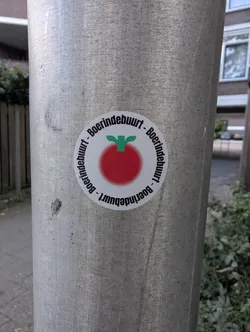 Street sticker A round white sticker on a metal post. It depicts a blurry red tomato-like shape with a green stem, bordered by the repeating black text 'Boerindebuurt'.