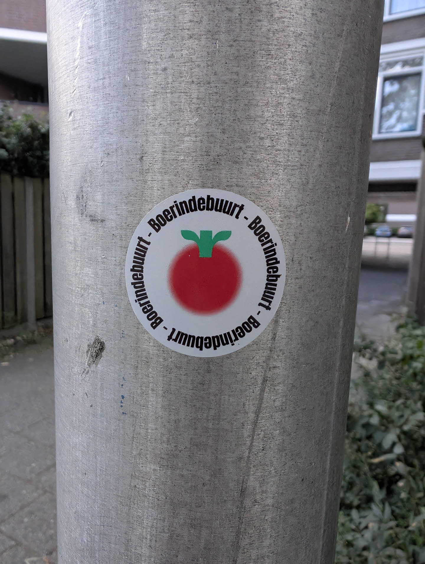 A round white sticker on a metal post. It depicts a blurry red tomato-like shape with a green stem, bordered by the repeating black text 'Boerindebuurt'.