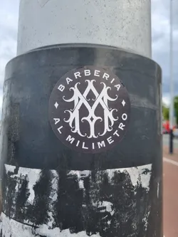 Street sticker A round black and white sticker featuring the logo of Barberia Al Milimetro. The logo is a monogram with stylized letters and ornate details. The text 'BARBERIA' and 'AL MILIMETRO' are written around the monogram. The sticker is affixed to a dark-grey pole.